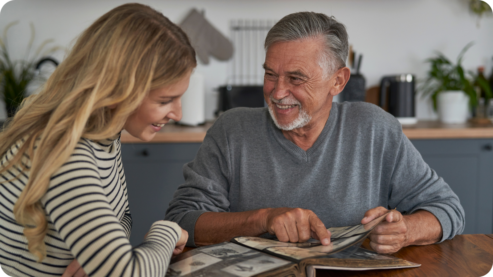 A parent and child sitting together at a table, gently turning the pages of a worn photo album while smiling at a tablet displaying the digitized images